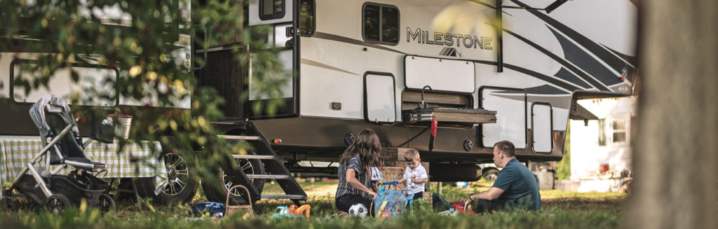 A family sitting in the grass outside an RV at a camp ground.