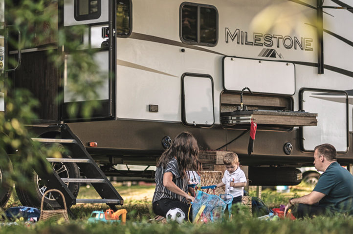 A family sitting in the grass outside an RV at a camp ground.