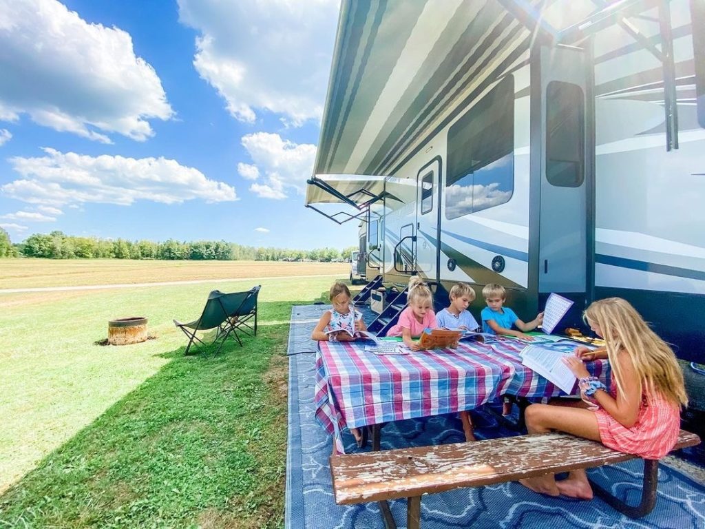 Four kids reading and doing homework at an outdoor picnic table next to an RV.