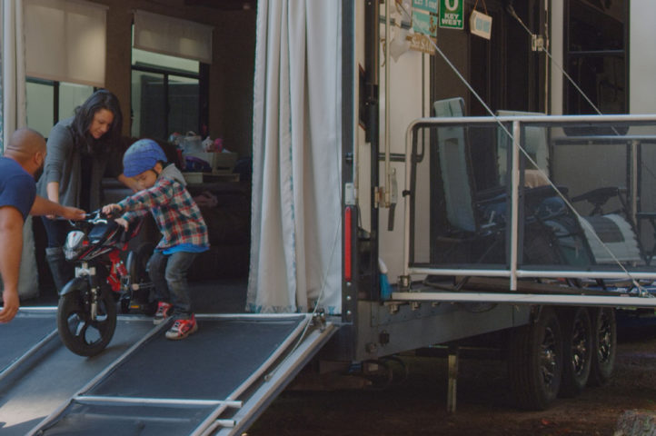 Parents helping a young kid walk a bicycle down a toy hauler ramp.