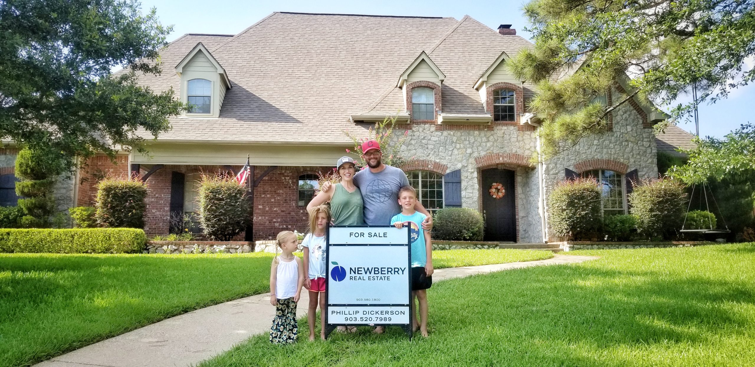 A smiling family behind a for-sale sign with a beautiful house in the background.