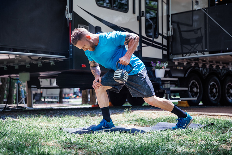 A fit man doing one-armed rows on a yoga mat outside his RV at a camp site. 
