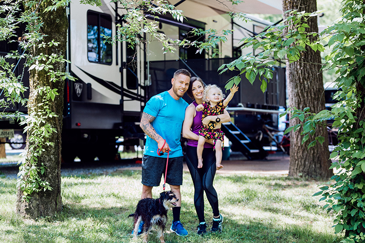 A young family holding a baby and standding with a dog on a leash, standing outside their RV at a campsite and smiling. 