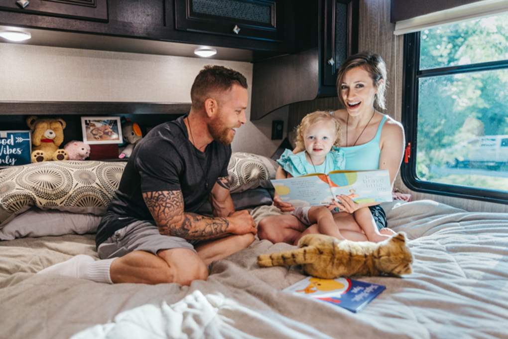 A young mom and dad reading a book to a little girl in an RV bedroom.