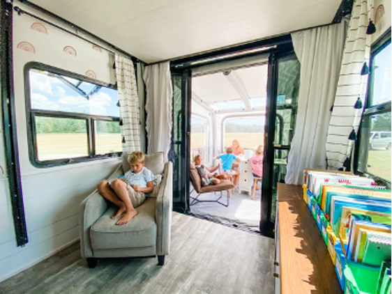 A view into multiple rooms of an RV, with a boy curled up in a chair reading a book and more kids on a patio beyond. 