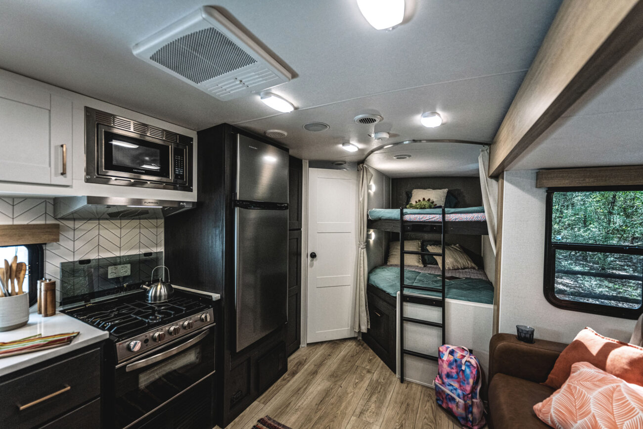 Interior of a travel trailer featuring a modern kitchen with an oven, stove, and microwave. Bunk beds with teal bedding are visible in the back. Theres a small sofa with orange cushions, a pink backpack on the floor, and a window with a view of trees.