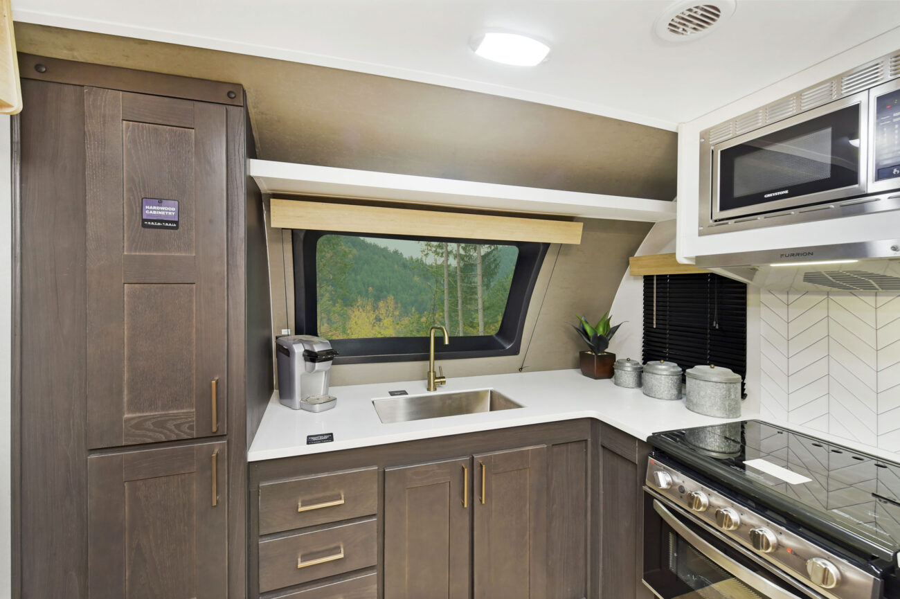 Modern RV kitchen with dark wood cabinets, white countertops, and a stainless steel sink. A window above the sink shows a forest view. There is a microwave, gas stove, and a coffee maker. Potted plant and containers are on the counter.