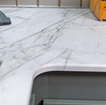 A close-up of a white marble kitchen countertop with gray veining, a corner of a sink, a folded gray towel, and part of a wooden item in the background.