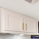 White overhead kitchen cabinets with gold handles, set against a white wall and ceiling. Part of a vent, windows, and a microwave are partially visible around the cabinets.