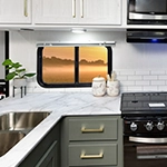 Modern kitchen with marble countertops, a stainless steel sink, a stove, and white cabinetry. A small potted plant and two jars sit on the counter near a window showing a sunrise or sunset.