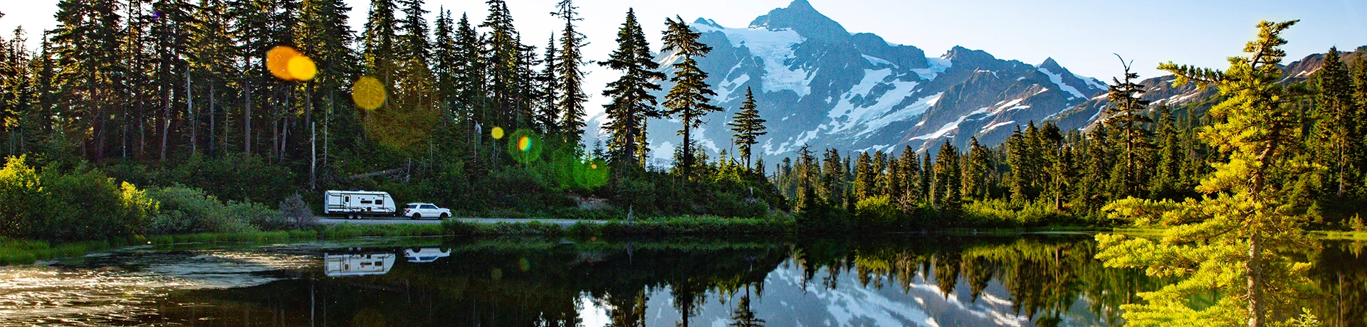 A white truck towing an Eddie Bauer Recreational Vehicle drives along a forested lakeside road, with tall pine trees and a snow-capped mountain reflecting in the calm water under a clear blue sky.