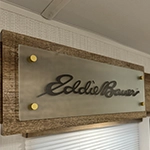 A metallic sign with the words Eddie Bauer in cursive script is mounted on a wooden panel above a window with a white blind.