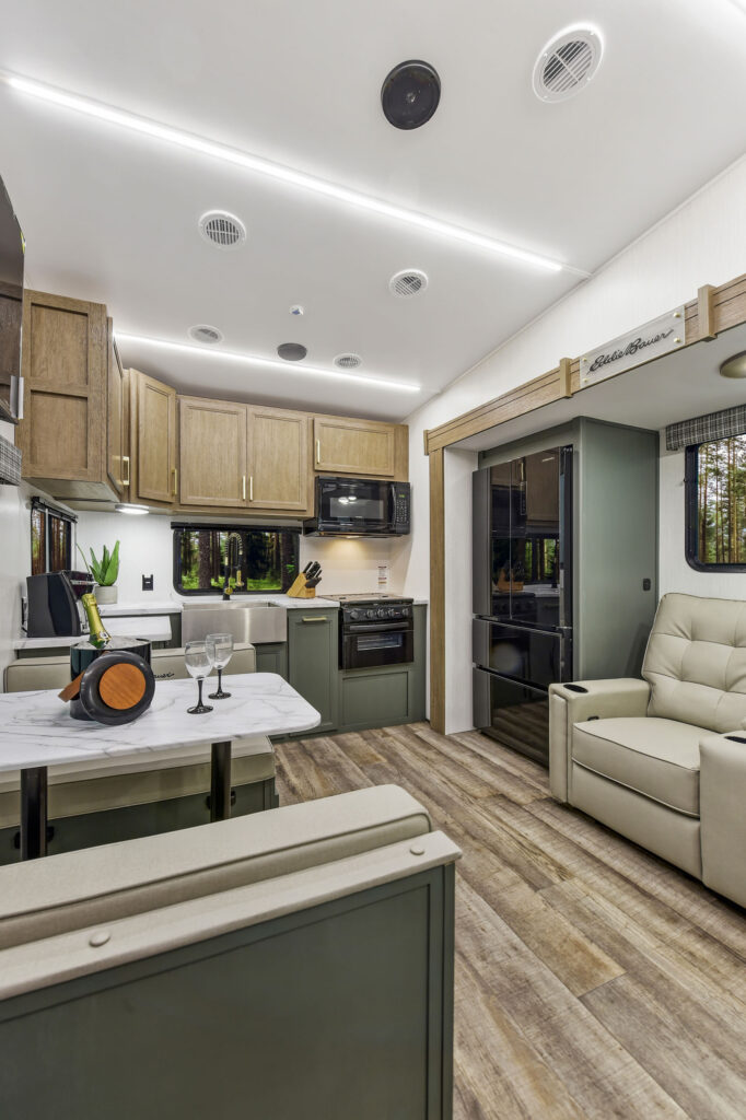 This image displays the 2025 Eddie Bauer EBF 2500RK travel trailer, featuring a beautifully designed rear kitchen layout. The kitchen boasts modern appliances, including a stove and refrigerator, along with generous counter space for meal preparation. Rich cabinetry and stylish backsplash add to the aesthetic appeal, creating a warm and inviting atmosphere. Adjacent to the kitchen, the open living area is furnished with comfortable seating, perfect for relaxation after a day of adventure. Large windows allow natural light to brighten the space, making it an ideal home away from home for your travels.