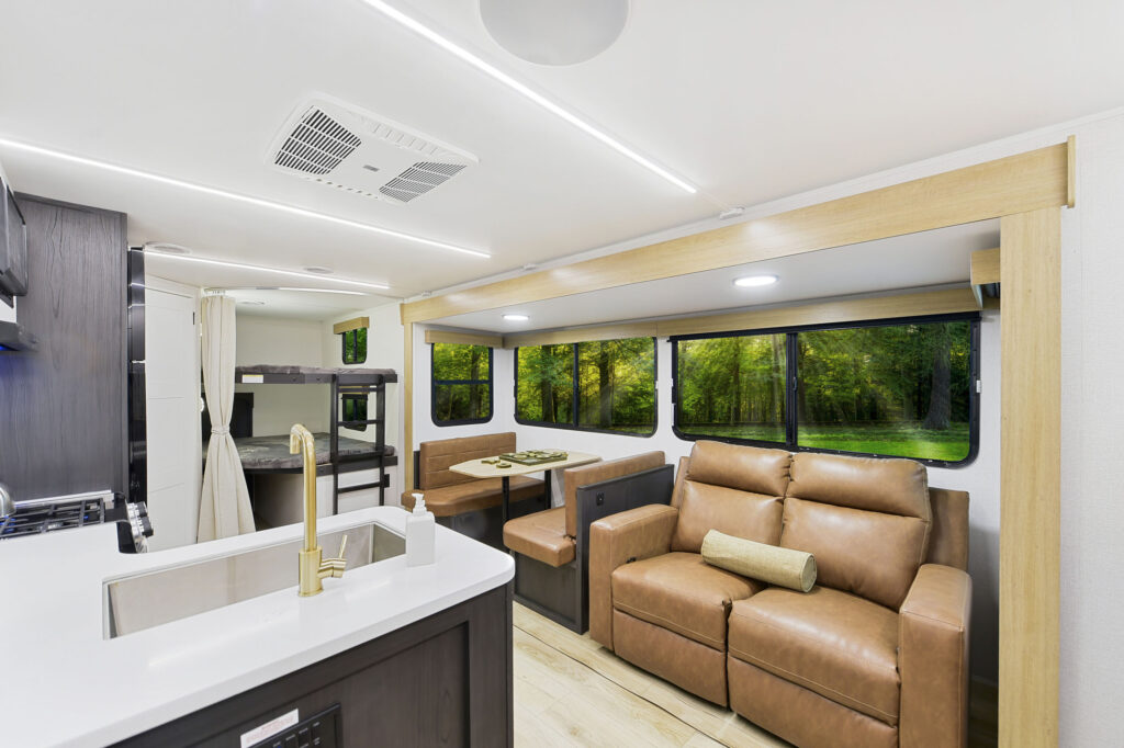 Interior of a modern RV featuring a kitchen with a sink, dining area with a table and bench seating, and a seating area with a brown leather couch. Large windows offer a view of a green forest outside.