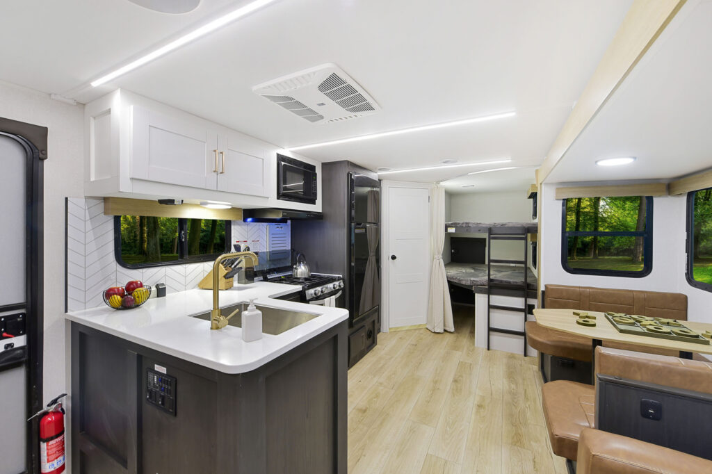 Interior of a modern RV featuring a kitchen with a sink and stove, a seating area with a table, bunk beds, and windows with scenic outdoor views. The space is well-lit with sleek, contemporary design elements and wood flooring.