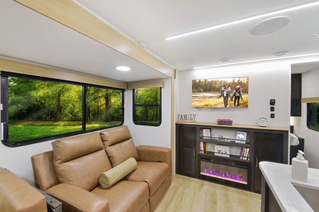 Interior of a modern RV living area with brown leather recliners, large windows showing a green forest, a small electric fireplace, and a TV above it. Decor includes a clock and FAMILY sign on the mantel.