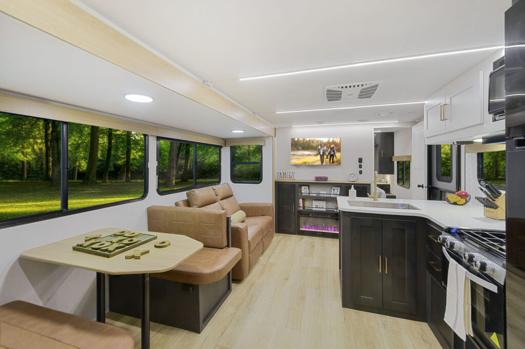 Interior of a modern RV featuring a kitchen with a stove and microwave, a seating area with a beige sofa, and a small dining table. Large windows offer views of a lush forest. The floor is light wood, and a digital fireplace is beneath a TV.
