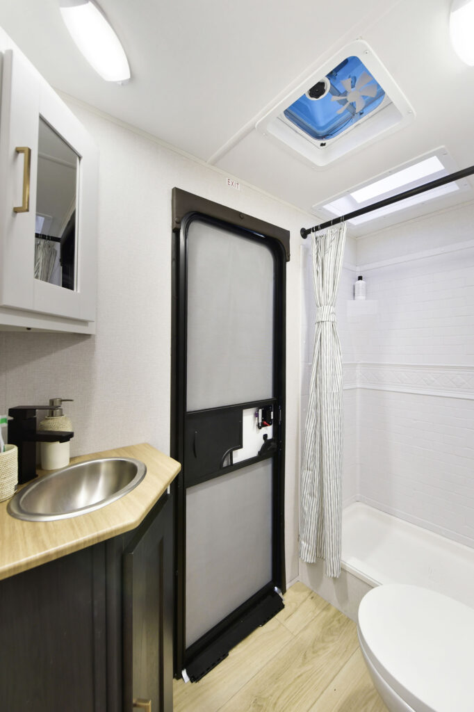 Interior of a compact bathroom featuring a small sink with a wooden countertop, a shower with a curtain, a white storage cabinet, and a ceiling vent. The floor is wooden.