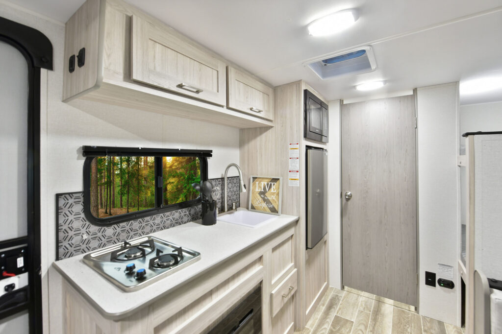 The image features the 2025 Prowler P17B travel trailer, showcasing its modern and compact design. The trailer boasts a sleek exterior with a contemporary color palette and stylish graphics, enhancing its appeal for outdoor enthusiasts. Large windows invite natural light into the living space while offering scenic views. The entryway is designed for easy access, leading into a thoughtfully arranged interior that maximizes space and comfort. With its blend of functionality and style, the P17B is perfect for weekend getaways and family adventures.