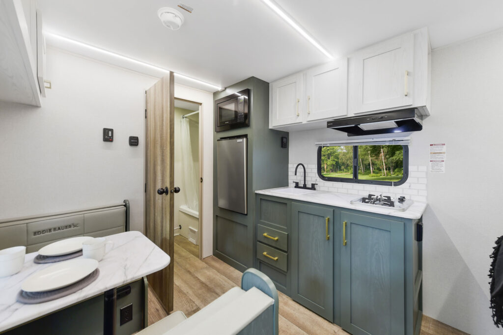 Modern RV interior with a dining area, kitchenette, green and white cabinets, stainless steel appliances, and a window above the sink showing greenery outside. A door leads to a small bathroom with a shower.
