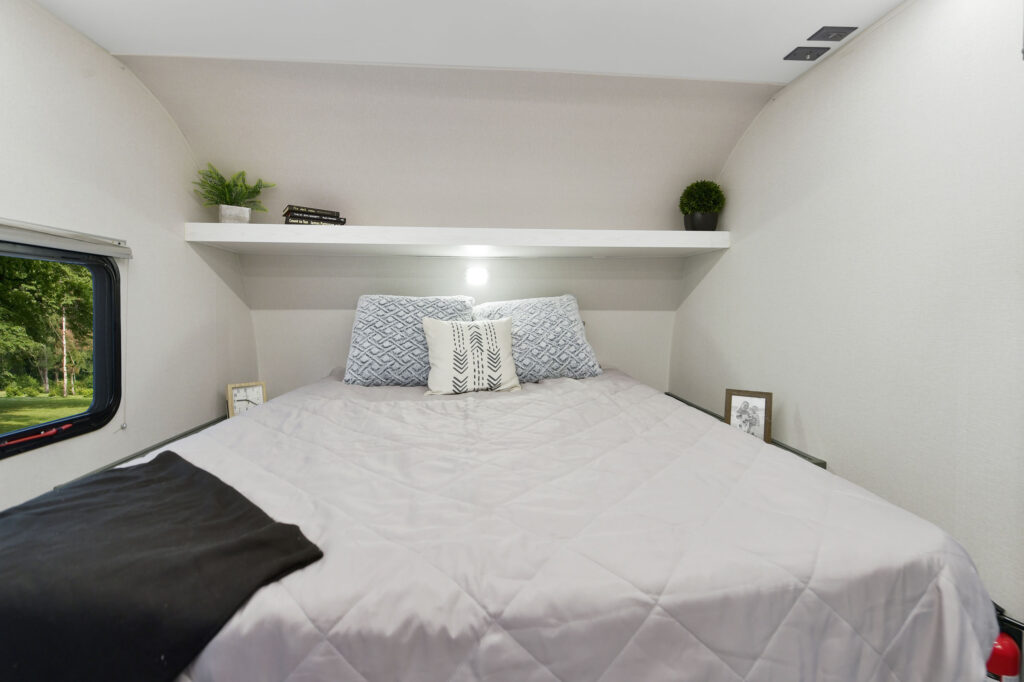 A neatly made bed with grey and white bedding in a compact, modern room. There are pillows, small framed photos, plants, and a decorative shelf above the bed. A window shows greenery outside.