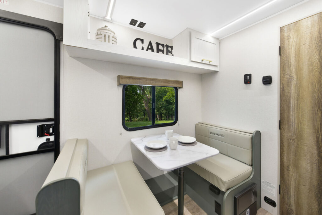 A modern RV dining area with a marble-look table, two cushioned benches, tableware set for two, and a window showing green trees outside. Overhead storage cabinets and a CAFE sign decorate the space.