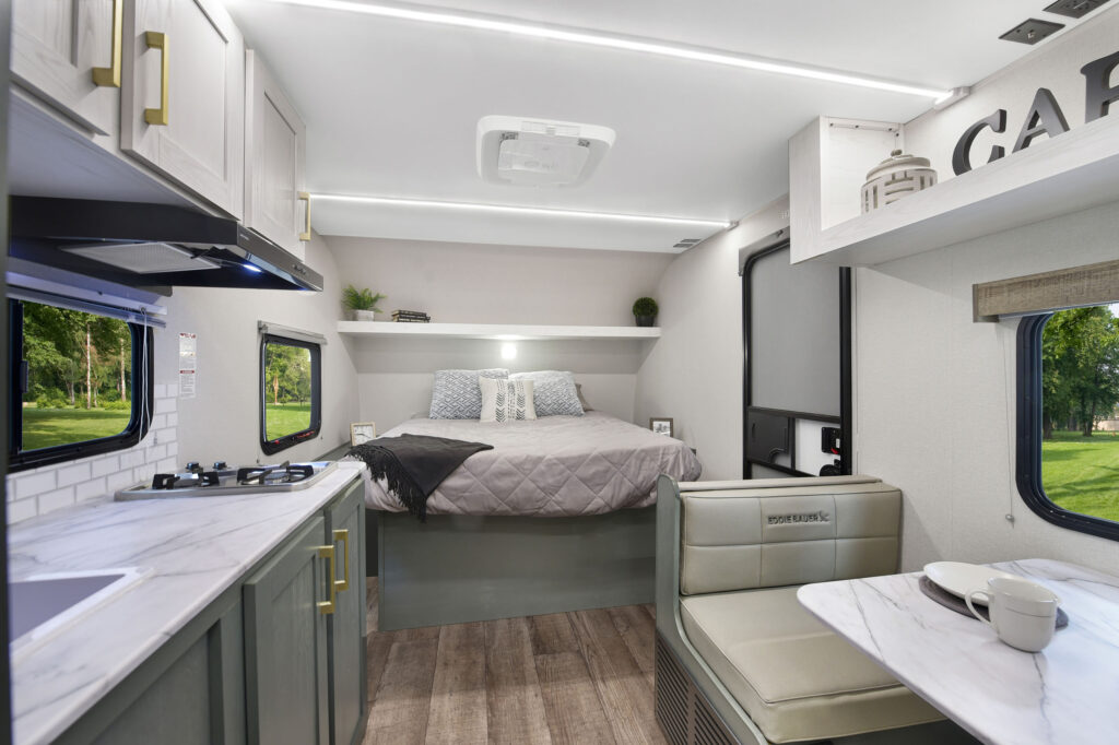 Modern RV interior with a cozy bed in the back, kitchen area with stove and sink on the left, dining booth on the right, and large windows letting in natural light. The decor is light and contemporary.