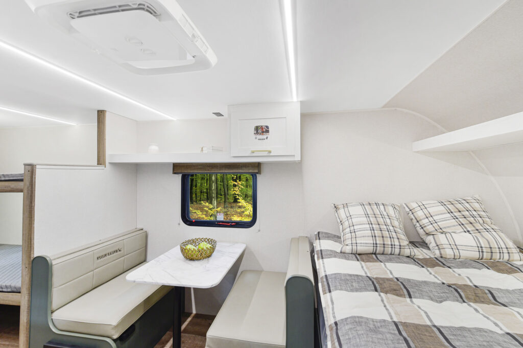 A cozy RV interior features a small dining area with a marble table and cushioned benches, a window with a forest view, built-in shelves, and a bed with plaid bedding. The space is bright with modern, minimal decor.