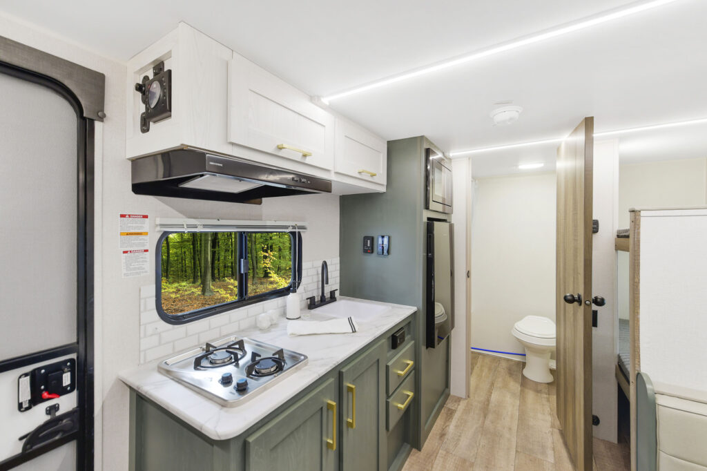 Modern RV kitchen with green cabinets, two-burner stove, sink, and window showing trees outside. A hallway leads to a bathroom with a toilet. Light wood flooring and overhead cabinets are also visible.