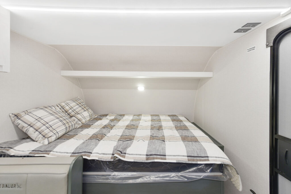 Interior of a small RV bedroom with a neatly made bed featuring plaid pillows and a striped comforter, a white shelf above the bed, and a wall light on a light-colored wall.