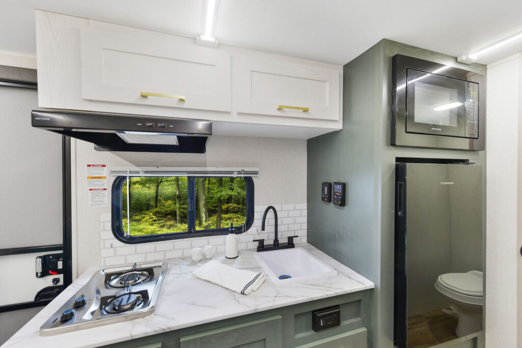 A compact RV kitchen with a marble countertop, two-burner stove, sink, white cabinets, a microwave, a small fridge, and a window showing a green forest outside. A towel and cup are on the counter.