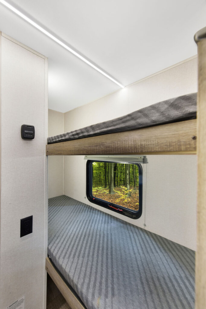 Two wooden bunk beds with gray-striped mattresses inside an RV or camper. A small window between the bunks shows a view of a green forest with sunlight filtering through the trees.