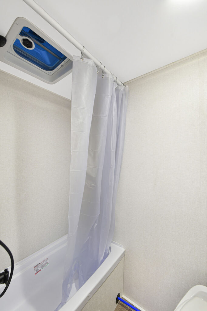 A small bathroom with a white bathtub, a translucent shower curtain partially closed, a vent with a blue cover in the ceiling, and beige textured walls.