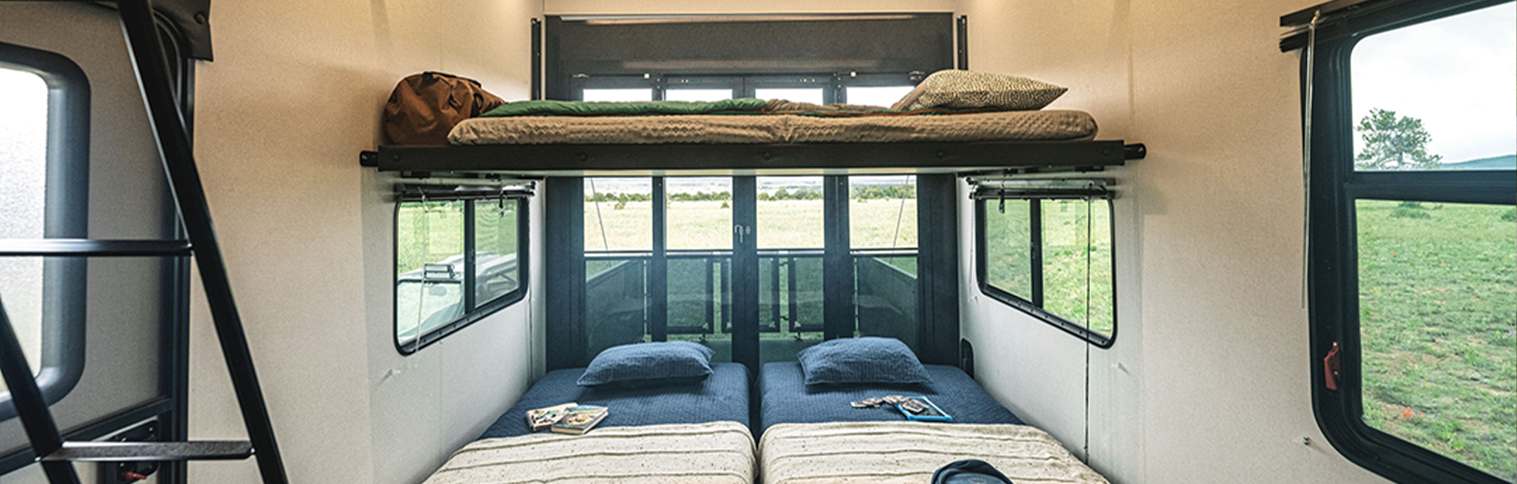 The interior of this 2025 RV toy hauler features a cozy sleeping area with twin beds below and a loft bed above. Large windows offer views of the greenery outside, while a ladder on the left provides easy access to the loft. Pillows and blankets are neatly arranged for comfort.