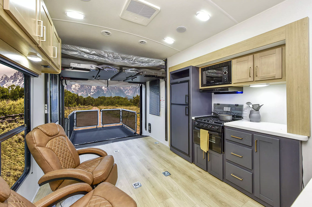 The interior of this 2025 toy hauler showcases a modern RV design, featuring an open seating area with two leather recliners. It includes a small kitchen equipped with a stove, microwave, and cabinets. An extended rear deck offers breathtaking mountain views through large windows, all within sleek and warm decor.