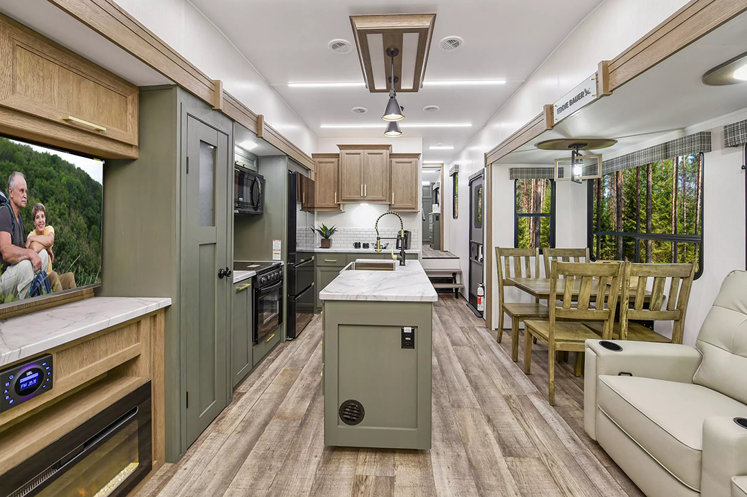 The interior of this 2025 model RV, reminiscent of luxurious fifth wheels, boasts a modern kitchen with a central island and sleek appliances. To the left, a fireplace rests beneath a TV, while the right hosts an elegant dining table. Wood and light tones complete the cozy seating area ambiance.