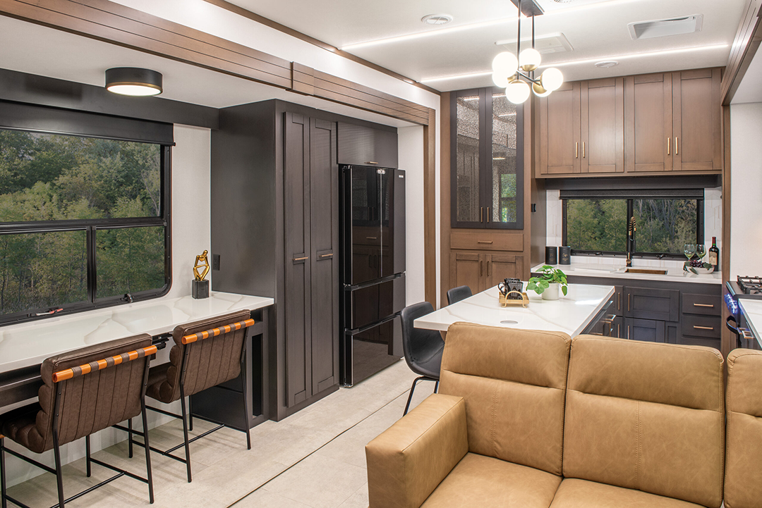 Step into this 2025 modern kitchen featuring wood cabinetry and a central island with a white countertop, complemented by sleek black appliances. Nearby, a brown sofa faces a dining table set by expansive windows, unveiling lush green views that could rival the comfort of any fifth wheel.