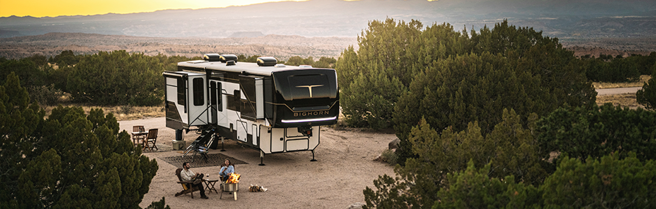 In a scenic desert area of 2025, a large RV is parked amid the trees. A family relaxes by a campfire, nestled into camping chairs with a table nearby. The sun sets in the distance, casting its warm glow over this serene landscape.