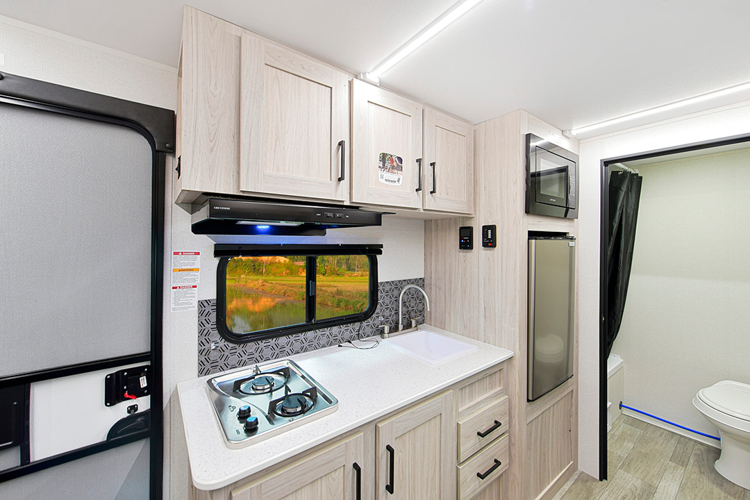 The 2025 RV kitchen perfectly suits modern families, featuring light wood cabinets, a two-burner stove, and a small sink. Below, drawers complement a compact refrigerator. The entrance to a small bathroom with a toilet is conveniently visible in the background.
