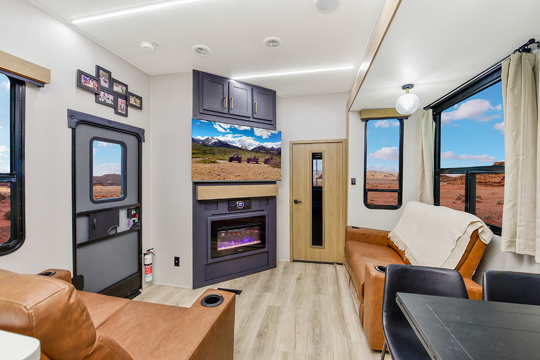 The 2025 interior of this modern RV, equipped as a multifunctional toy hauler, features a cozy living area with a brown sofa, armchair, and wall-mounted TV above an electric fireplace. Large windows unveil desert vistas, while light wood flooring and decor enhance the warm ambiance.