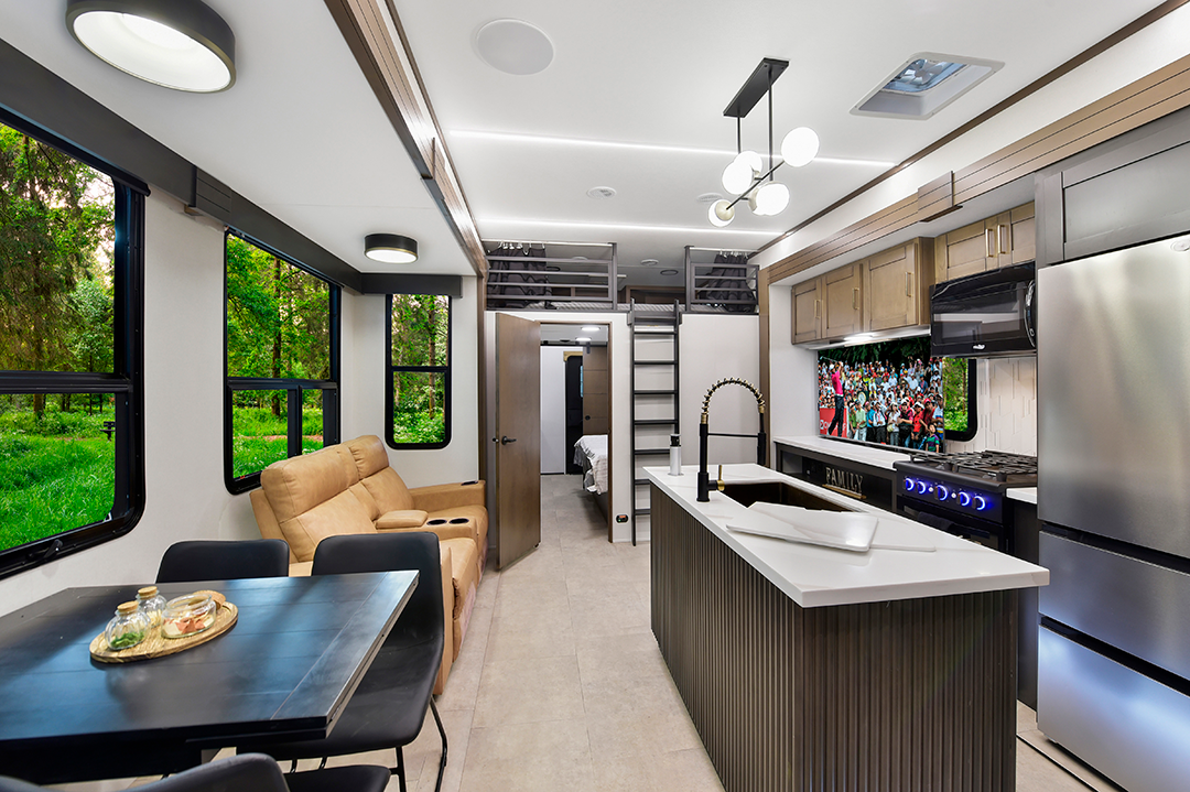 The interior of a modern RV, designed with 2025 families in mind, features a kitchen with stainless steel appliances, a dining table for cozy meals, and a recliner facing the TV. Large windows invite lush greenery inside, while a loft and closed bedroom door offer both space and privacy.