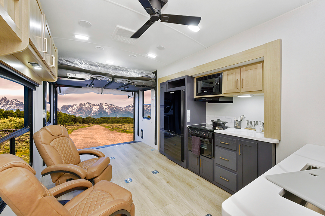 Inside the 2025 RV toy hauler, youll find two tan chairs, a sleek kitchenette, and a ceiling fan. The open rear door frames a scenic view of a dirt road stretching toward snow-capped mountains and lush green hills.