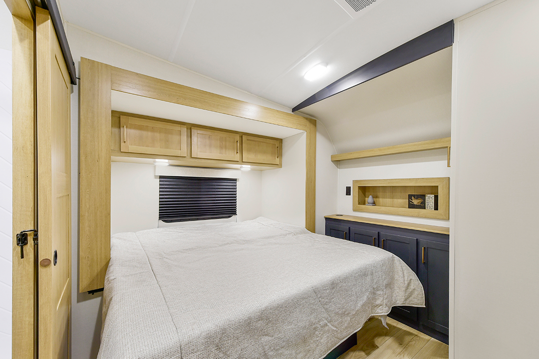 The interior of this 2025 toy hauler features a cozy bedroom with a double bed adorned in light-colored bedding. Overhead cabinets sit above the bed, complemented by dark cabinetry on the right. Crisp white walls and wooden flooring add warmth, while a small window with a dark blind rests above the bed.