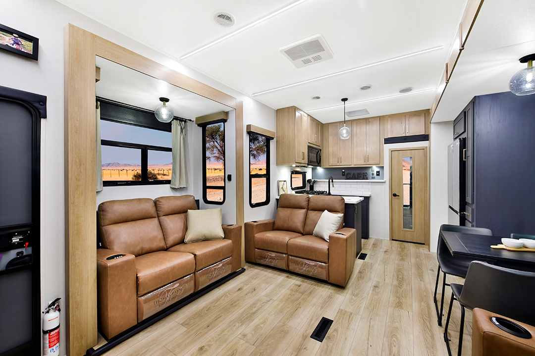 The interior of this stylish 2025 RV boasts a modern design with two brown leather sofas, wooden flooring, and a kitchenette featuring light wood cabinets. A small dining area complements the space, while large windows in the toy hauler reveal stunning views of the desert landscape outside.