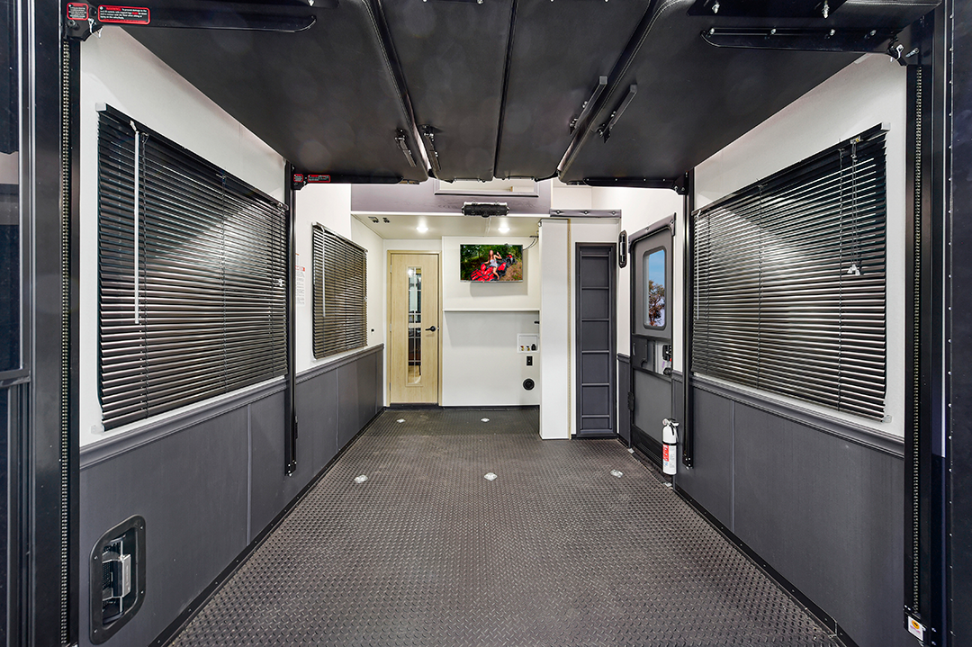 Inside the 2025 toy hauler, discover a spacious cargo area with textured flooring. Closed blinds cover the side windows, and a rear door opens to a warmly lit interior room. A small fire extinguisher is conveniently mounted on the wall for safety.