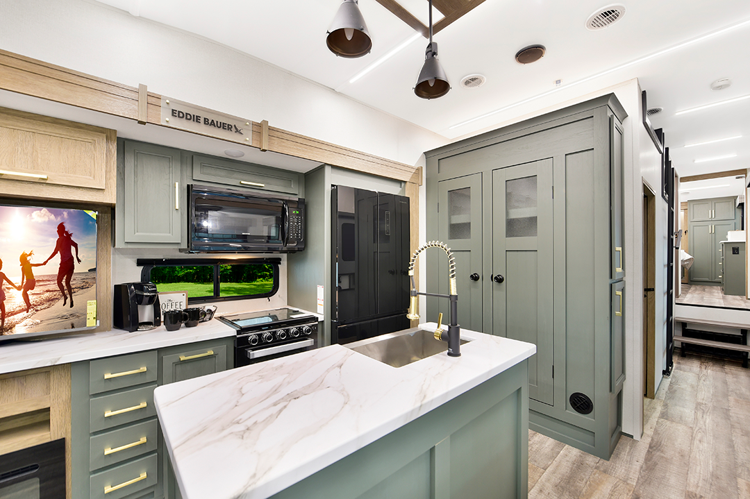 The interior of this modern 2025 RV kitchen, reminiscent of luxurious Fifth Wheels, showcases green cabinetry and a marble countertop island. Complete with a stove, microwave, and sink, its decor includes a scenic photograph and potted plant. Natural light beautifully illuminates the space.
