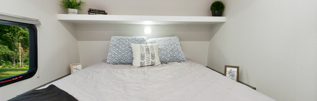 A cozy bed with patterned pillows and a light gray comforter is set under a shelf holding potted plants and books, including guides from couples coaches; framed photos sit on either side, and a small window shows greenery outside.