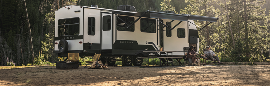 A modern, innovative travel trailer with an extended awning is parked at a campsite surrounded by trees. Two people relax in chairs by a campfire, enjoying the outdoors and cool shade.