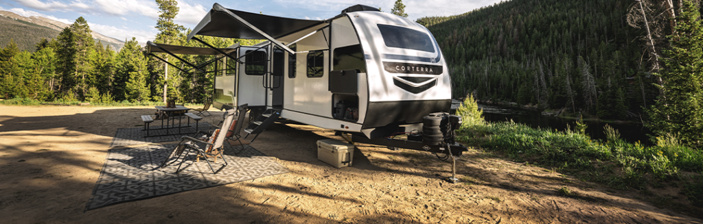 A modern white travel trailer with auto-leveling and awnings extended is parked at a scenic campsite near a river, surrounded by lush green forest and mountains. Outdoor chairs and a table are set up on a patterned mat beside the trailer.