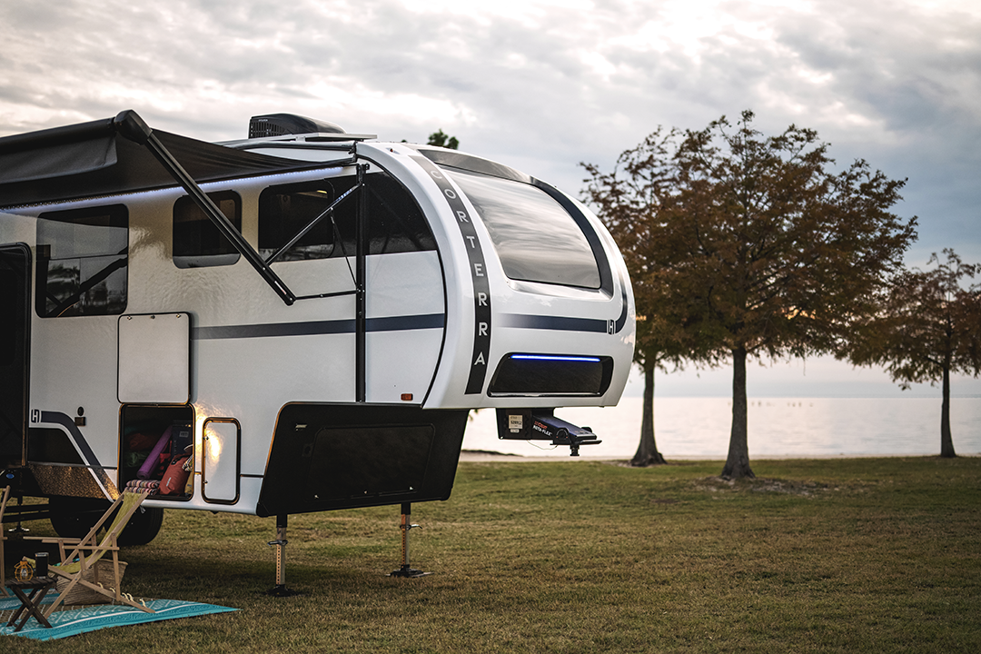 A modern white camper trailer with auto-leveling is parked on grassy ground near a lake, with trees in the background and cloudy skies overhead. An awning is extended, and a camping chair sits outside the open door.